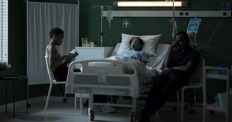 Young African mother reading a book father sitting on a chair by the bed in a hospital where a child is unconscious.