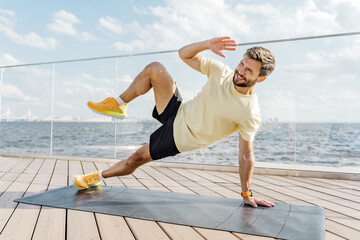 Man performs side plank exercise on yoga mat by the water during a sunny day
