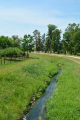 A stream flows through a lush green meadow in Erdody Park, Jastrebarsko, Croatia, under a bright blue sky with a path and trees lining the edges of the park