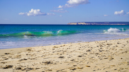 A beautiful beach with soft, white sand and crystal-clear turquoise water. Gentle waves lap at the shore, creating a tranquil atmosphere. In the distance, a rocky headland rises from the sea.

