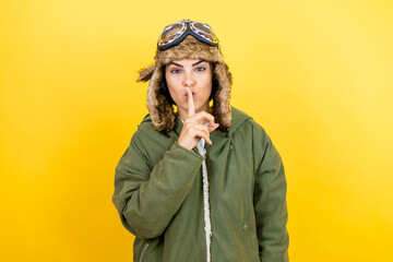 Young beautiful aviator woman wearing vintage pilot helmet with glasses and jacket over yellow background asking to be quiet with finger on lips. Silence and secret concept.