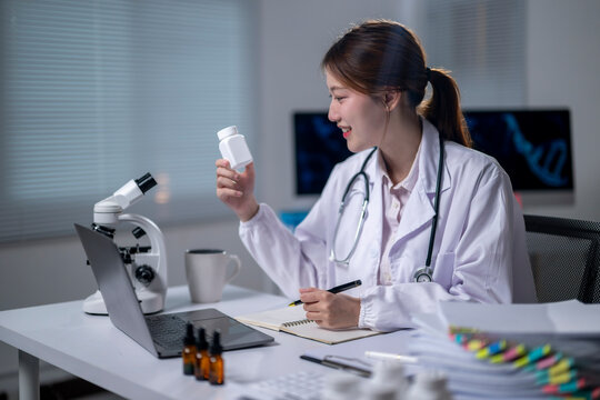 A woman in a white lab coat is sitting at a desk with a laptop and a notebook