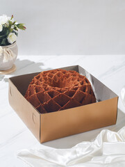A beautifully designed bundt cake sits in a simple brown box, accompanied by fresh flowers and elegant fabric on a light setting
