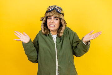 Young beautiful aviator woman wearing vintage pilot helmet with glasses and jacket over yellow background clueless and confused expression with arms and hands raised