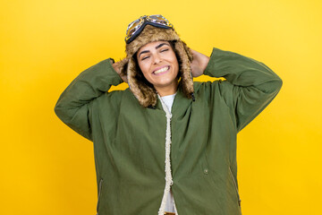 Young beautiful aviator woman wearing vintage pilot helmet with glasses and jacket over yellow background relaxing and stretching, arms and hands behind head and neck smiling happy