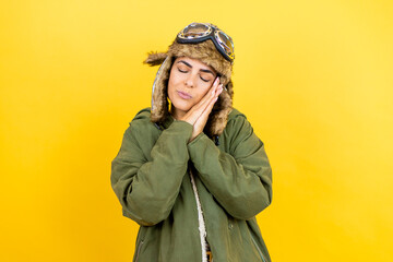 Young beautiful aviator woman wearing vintage pilot helmet with glasses and jacket over yellow background sleeping tired dreaming and posing with hands together while smiling with closed eyes.