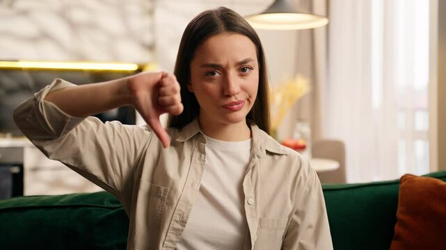 Upset frown frustrated dissatisfied Caucasian female lady girl woman indoors in living room at home at couch showing thumb down gesture dislike hand sign bad negative result failure looking at camera
