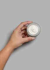 Female hand holding opened cream jar over grey background top view, negative space