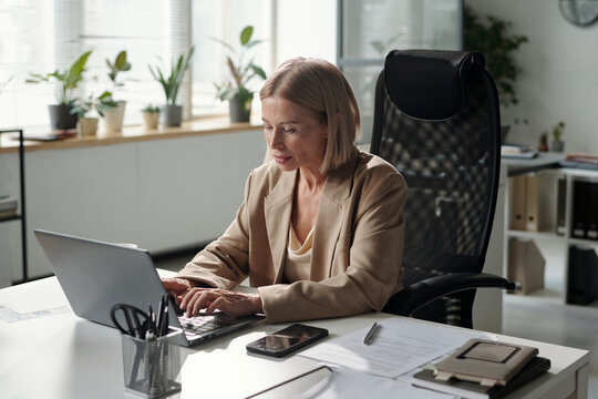 Serious mature blond chief executive officer in formalwear typing on laptop keypad by workplace in office - Powered by Adobe