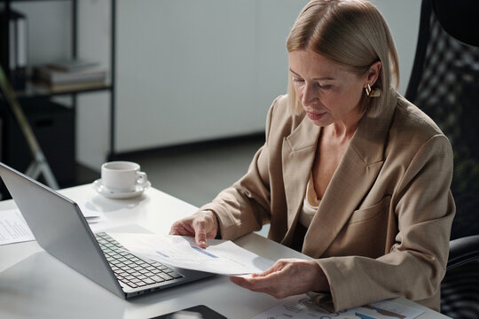 Blond businesswoman in elegant beige suit looking through graphic data or contract terms on financial document