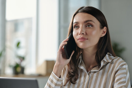 Young brunette female white collar worker speaking on smartphone while sitting by workplace in front of camera