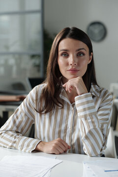 Confident female office manager looking at camera while sitting by workplace and touching her chin