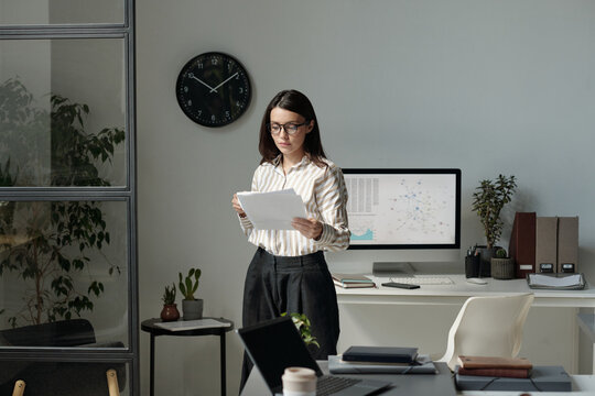 Serious female accountant in smart casualwear and eyeglasses standing by workplace and reading paper
