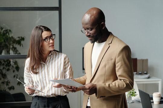 Young confident businesswoman looking at male colleague reading financial paper at working meeting
