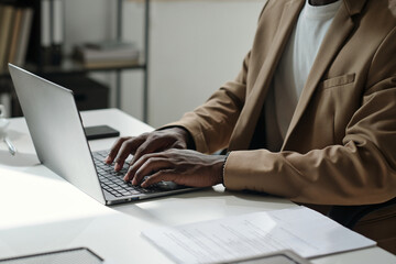 Hands of young African American male manager in formalwear sitting by desk and typing on laptop keypad