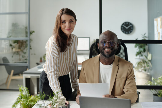 Two happy young intercultural economists looking at camera while woman standing next to man sitting by desk