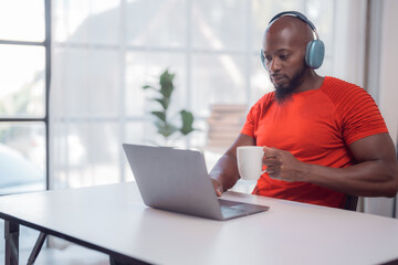 Young black professional wearing headphones is working on his laptop while enjoying a cup of coffee, showcasing the modern work-from-home lifestyle