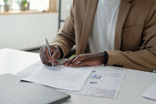 Hands of young unrecognizable male economist with pen reading business contract and signing it by desk