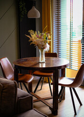 A table with four chairs and a vase of flowers in the center. The table is surrounded by a couch and a window