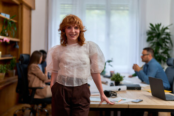 Redhead businesswoman is posing in a modern office with hands in pockets while her team is working in the background, demonstrating leadership and teamwork in a collaborative workspace
