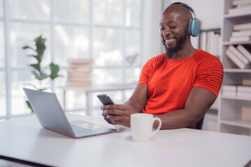 Happy african american man wearing headphones using smartphone and laptop while sitting at desk, working remotely from home and enjoying music