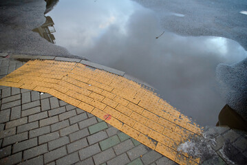 Large puddle on city street reflecting the cloudy sky near worn yellow tactile paving designed for...