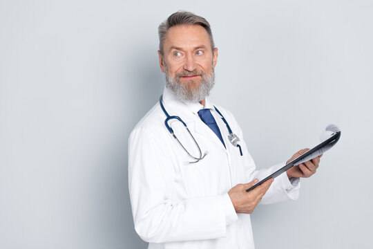 Male doctor in white coat smiling while holding clipboard against a light grey background in a professional medical studio setting
