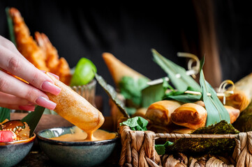 A close-up of a hand dipping a crispy spring roll into a flavorful sauce. In the background, a variety of appetizers are arranged artistically with fresh greens and garnishes.