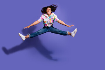 Obraz premium Young woman with curly hair joyfully leaps in mid-air on a vibrant purple background, showcasing colorful casual attire and white sneakers