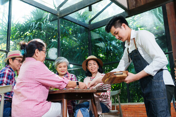 Senior Asian woman with close friend after retirement being served by a waiter at cafe. Elderly friends enjoying delicious treats, engaged in conversation, and sharing joyful moments together.