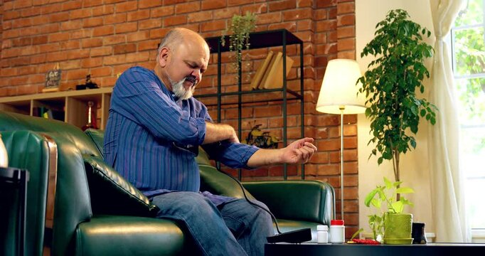 Indian senior man checks blood pressure at home with a digital monitor, sees normal reading, and smiles while holding a pill bottle for routine health care, sitting on sofa in modern living room