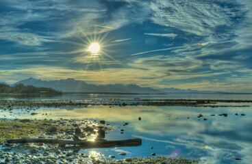 Ein Sonnenstern bei blauem Himmel mit kleinen Wolken über dem Alpenpanorama am Chiemsee mit Spiegelung 