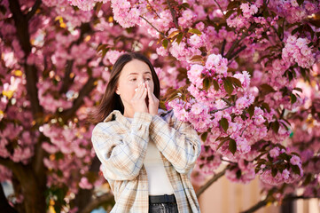 Woman allergic suffering from seasonal allergy at spring in blossoming garden at springtime. Woman sneezing and blowing nose using nasal handkerchief in front of blooming tree. Spring allergy concept.