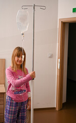 Little girl receiving intravenous therapy, holding a drip stand in a hospital room, recovering from...