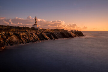A picturesque lighthouse stands on a rocky cliff during sunset in Menorca Spain. The dramatic sky...