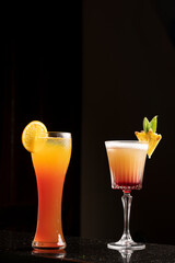 Glass of orange juice with slice garnish and a pina colada cocktail placed side by side, styled on a black background for a bold and vibrant tropical beverage presentation.
