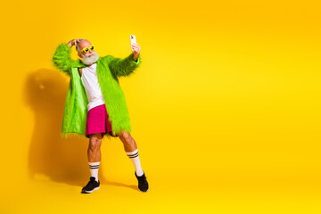 Elderly man in vibrant green fur coat taking a selfie against yellow background, expressing unique style, confidence, and freedom
