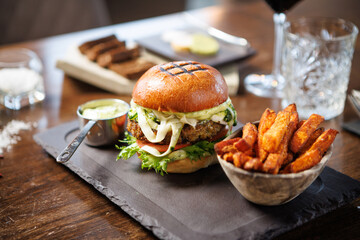 Vegetarian burger with fresh greens, tomato, cheese, and creamy sauce on a brioche bun. Accompanied by a bowl of sweet potato fries and a metal cup of dip. Healthy and tasty dining option
