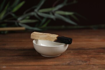 Burnt palo santo stick and bowl on wooden table against black background, closeup