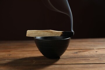 Smoldering palo santo stick and bowl on wooden table against black background, closeup