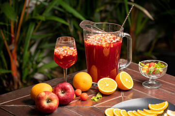 Sangria served in a glass jug and wine glass on a table, with whole and sliced fruits like oranges...