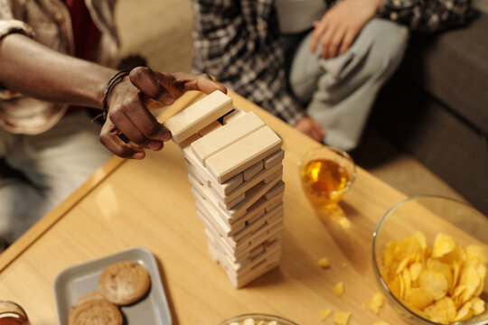 High angle of hand of young man putting or taking wooden block from top of tower built on table during game