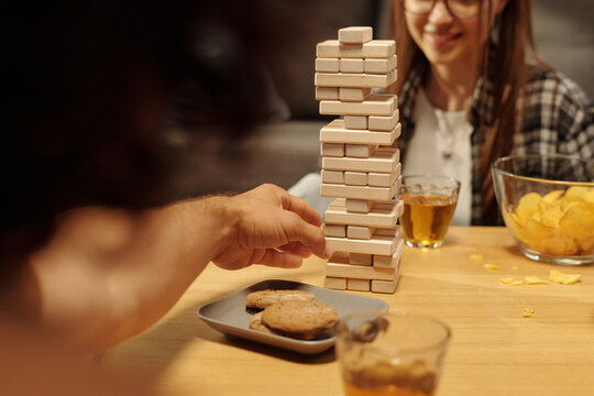Hand of guy taking block out of wooden tower stack during tabletop game with his girlfriend at leisure