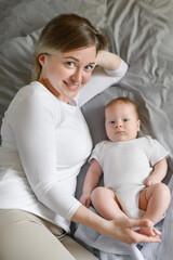 Sweet mom lies on bed next to baby, lovingly holding his legs, looking with tenderness and love. Concept of family, love and care for baby, happy motherhood. Vertical.