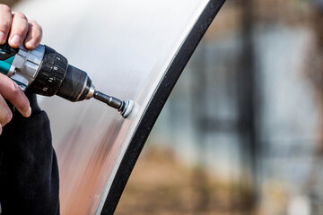 A man holds a battery-powered screwdriver and assembles a new greenhouse made of polyethylene,...