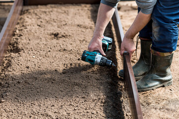 A man holds a battery-powered screwdriver and collects new beds from WPC into a greenhouse made of polyethylene, polycarbonate. Macro photography. Side view. Preparing for the spring garden season.