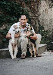 A man in affectionately embraces two dogs. One of the dogs , while the other is brown and short fur. The scene reflects a moment of connection and affection between the man and his pets.