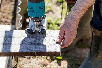 A man holds a battery-powered electric saw and builds new beds in a greenhouse made of wood-polymer...