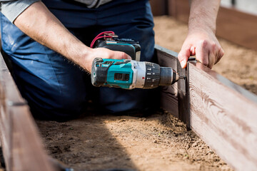 A man holds a battery-powered screwdriver and collects new beds from WPC into a greenhouse made of polyethylene, polycarbonate. Macro photography. Side view. Preparing for the spring garden season.