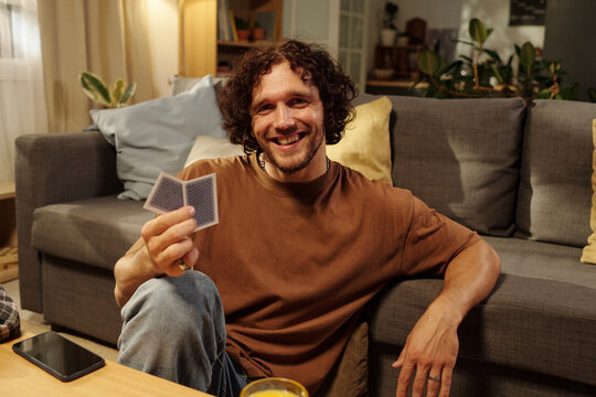 Young laughing man with two playing cards in hand looking at camera while sitting by couch on the floor